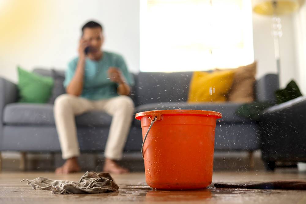 Roof leak dripping water into bucket in living room of home. A man in the background is on the phone.