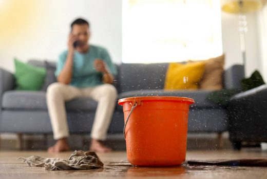 Roof leak dripping water into bucket in living room of home. A man in the background is on the phone.